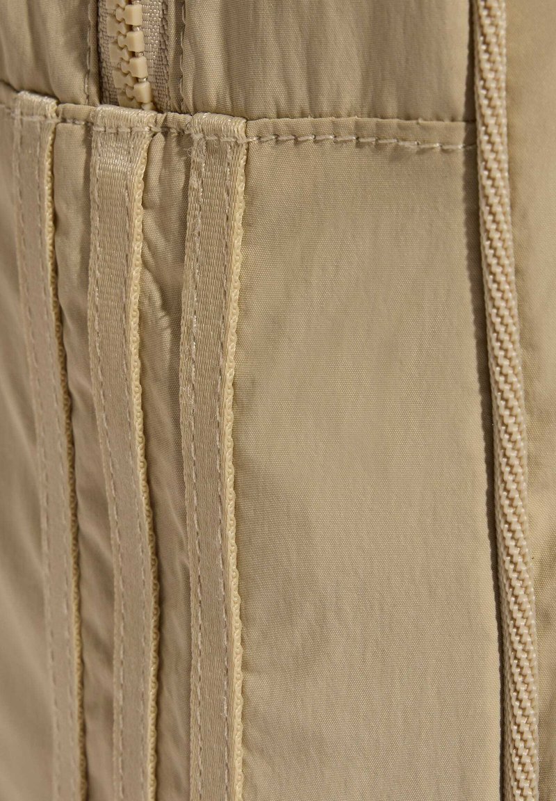 Primo piano di tessuto beige con cuciture verticali, cinturini sovrapposti e dettaglio con cerniera in plastica sul lato sinistro.