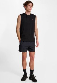 Svart ärmlös tröja med liten röd logga, kombinerad med marinblå shorts. Modellen bär svarta strumpor och sneakers, står mot en vit bakgrund.