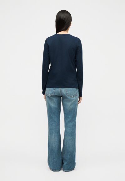 Γυναίκα που στέκεται γυρισμένη με την πλάτη, φοράει μπλε σκούρο μακρυμάνικο πουκάμισο και ανοιχτό μπλε τζιν καμπάνα, μπροστά σε απλό λευκό φόντο.