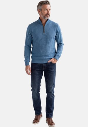 Mężczyzna z siwymi włosami i brodą, ubrany w jasnoniebieski sweter zapinany na zamek do połowy, ciemne dżinsy i brązowe trampki, stojący z jedną ręką w kieszeni.