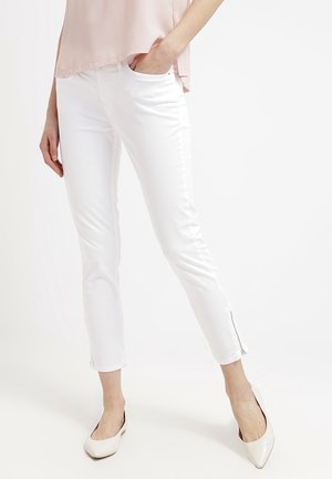 Frau trägt weiße, eng anliegende Knöchellange Hosen mit seitlichem Reißverschluss und hellrosa Oberteil, steht mit einer Hand in der Tasche und weißen spitzen Ballerinas.