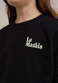 Fekete pulóver puha anyagból, a mellkason könnyű zöld "Le Marais" logóval, játékos betűtípussal. Kerek nyakkivágás.