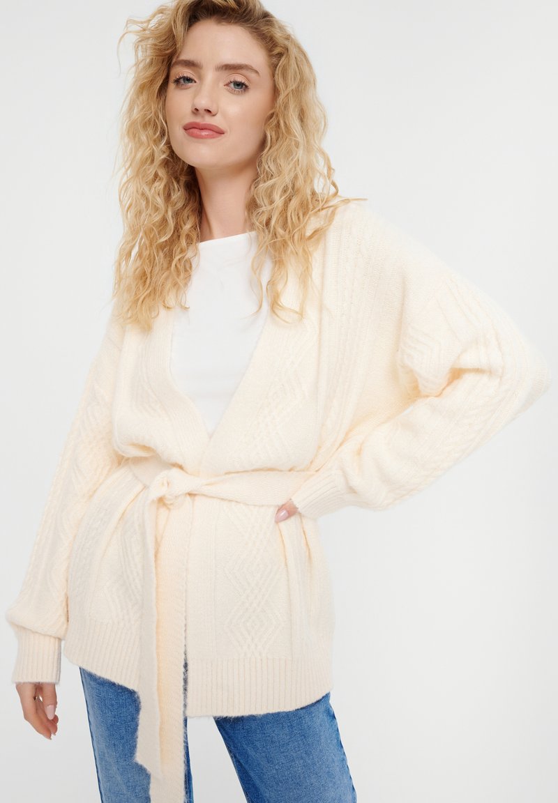 Cremfarbener gestrickter Cardigan mit einem strukturierten Rautenmuster, langen Ärmeln und einem gebundenen Saum. Kombiniert mit einem schlichten weißen Shirt und Jeans.