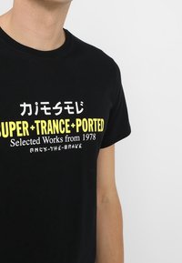 Musta puuvillapaita, jossa on rohkeaa keltaista tekstiä "SUPER + TRANCE + PORTED" sekä lisä Japanilaisia merkkejä. Lyhyet hihat, klassinen malli.