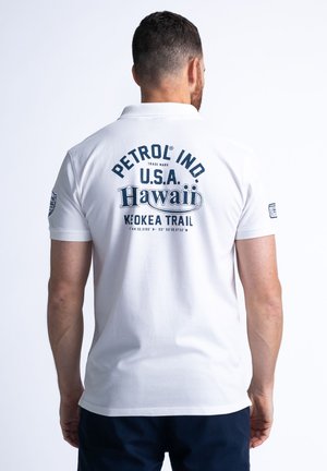 Witte polo shirt met een marineblauw grafisch print op de achterkant, met de tekst "PETROL IND. U.S.A. Hawaii Keokea Trail," en een klassieke kraagontwerp.