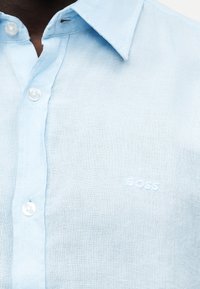 Gros plan d’une chemise bleue pâle à boutons, avec col et « BOSS » brodé en fils blancs sur la poitrine.