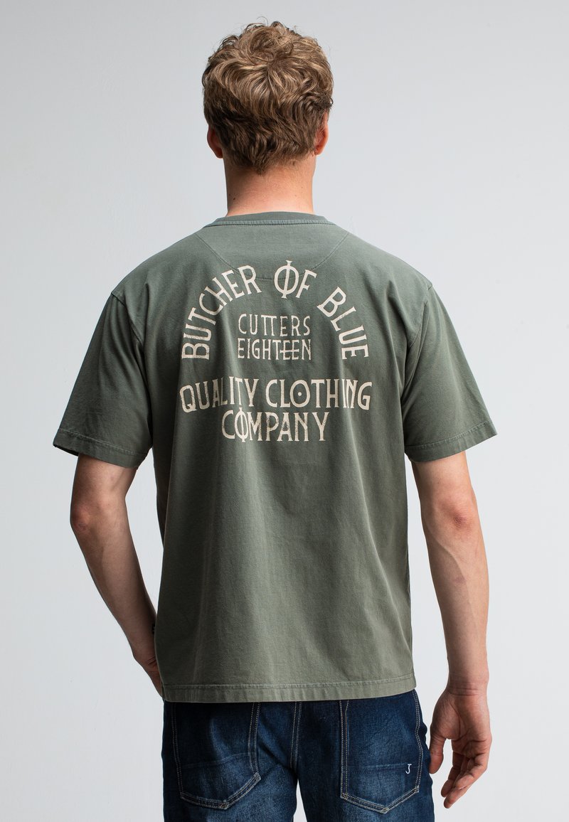Man met kort krullend haar, gekleed in een olijfgroen T-shirt met de tekst "Butcher of Blue Quality Clothing Company" op de rug, gecombineerd met een blauwe spijkerbroek.