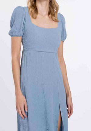 Femme portant une robe bleu clair texturée avec des manches bouffantes, un décolleté carré et une fente sur le côté droit.
