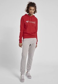 Röd huvtröja med vit logotyp, grå mjukisbyxor och gråa sneakers. Mjukt material, avslappnad passform och elastiska muddar. Avslappnad sportklädsel.