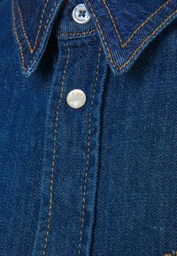 Koszula dżinsowa w ciemnym niebieskim kolorze, z ostro zakończonym kołnierzem, perłowymi napami oraz kontrastującym złotym przeszyciem wzdłuż szwów.