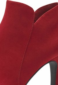 Stivaletto alla caviglia in suede rosso con punta affusolata e una scollatura curva lungo la parte superiore. Presenta un tacco a stiletto nero. Texture complessivamente liscia.