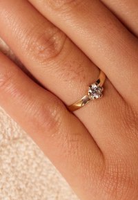 Gold ring with a round, clear gemstone center; smooth band features a polished finish; worn on a hand with textured skin.