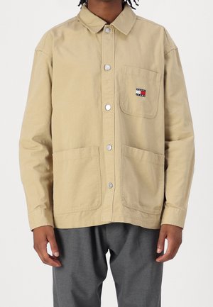 Osoba nosząca beżową kurtkę zapinaną na guziki z trzema dużymi kieszeniami, jedna z nich zdobi małe logo Tommy Jeans, w zestawie z szarymi spodniami.