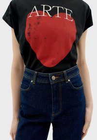 Zwarte katoenen t-shirt met een grote rode hartafbeelding en witte tekst "ARTE," gecombineerd met hoge donkere blauwe spijkerbroek.