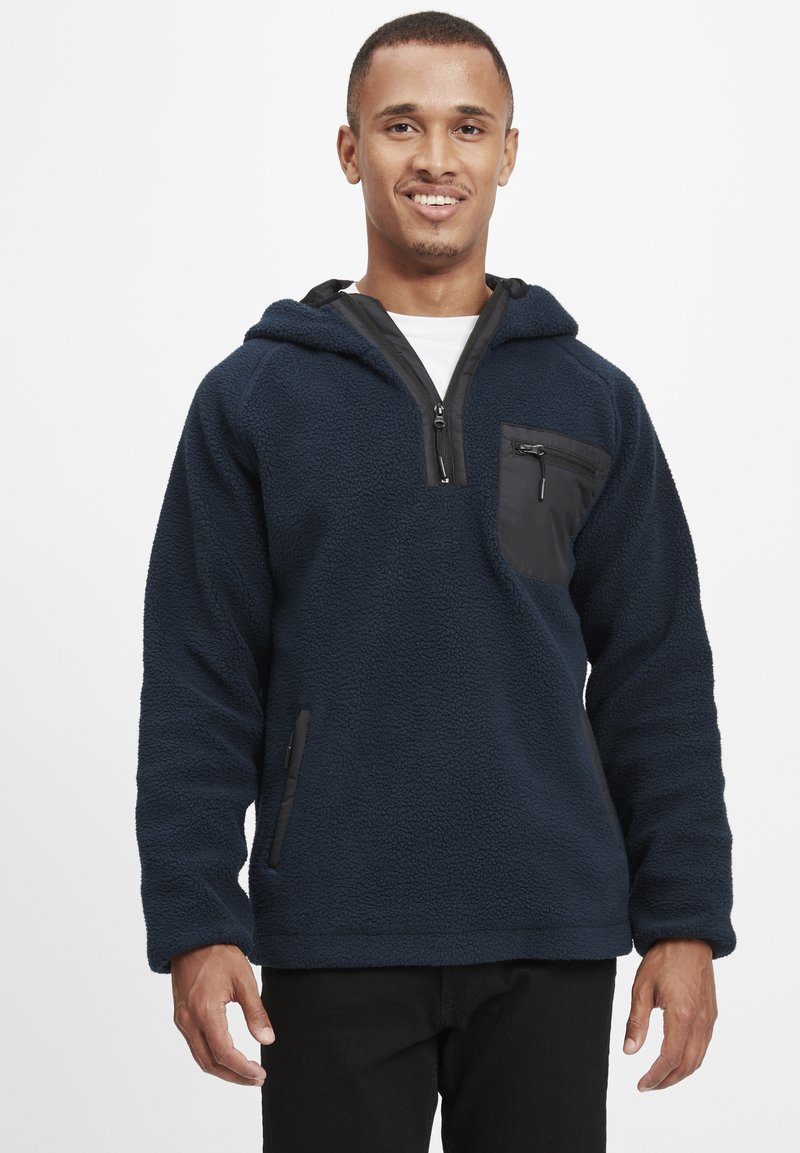 Marineblå fleece pullover hoodie med en sort lynlåslomme på brystet, sidelommer og et tekstureret stof for varme.