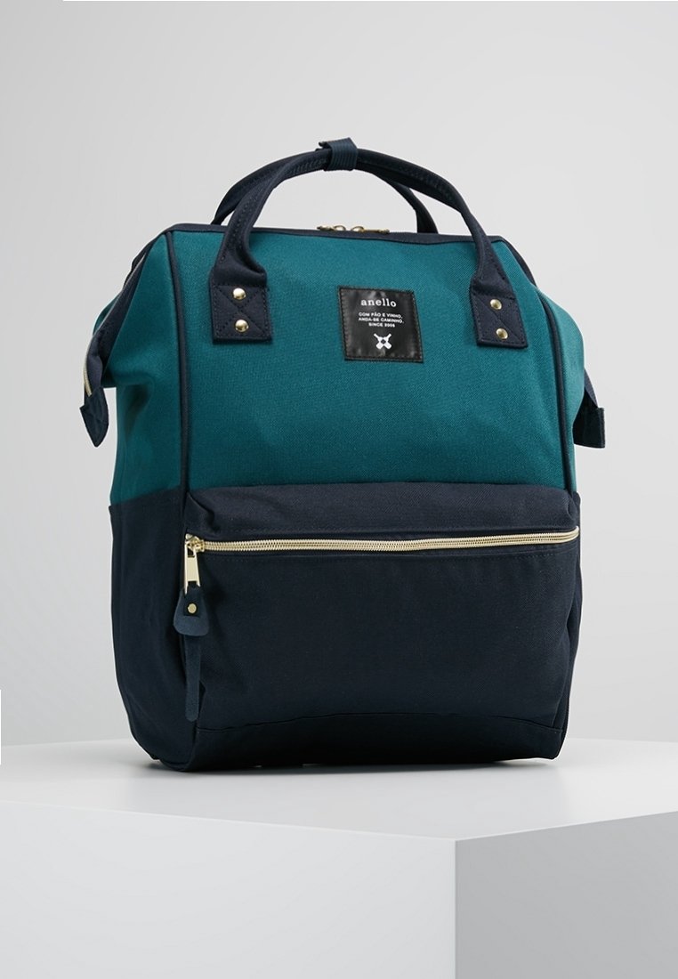 Sac à dos rectangulaire bleu sarcelle et noir avec poignées supérieures, poche avant zippée et petite étiquette de marque sur le panneau avant supérieur.