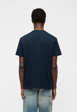 Muž s krátkymi kučeravými vlasmi otočený chrbtom, oblečený v obyčajnej tmavomodrej košeli s krátkym rukávom a svetlomodrých rifliach s zadnými vreckami.