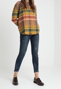 Rutig skjorta i gult, orange och grönt, kombinerad med mörkblå croppade jeans med slitna kanter och bruna snörskor.