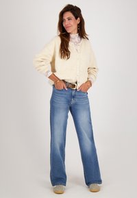 Crème cardigan met knoopsluiting, wijde mouwen, gedragen over een kanten top. Hoog geplaatste blauwe jeans met een relaxte pasvorm en een lichte vervaging. Casual schoenen.