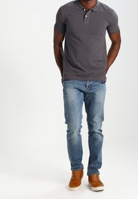 Man som bär en mörkgrå pikétröja, ljusblå jeans och bruna högskor som står med händerna bakom ryggen.