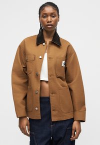 Kvinne med flettet hår iført en brun knappet jakke med svart krage, hvit cropped topp og mørkeblå jeans mot en ensfarget bakgrunn.