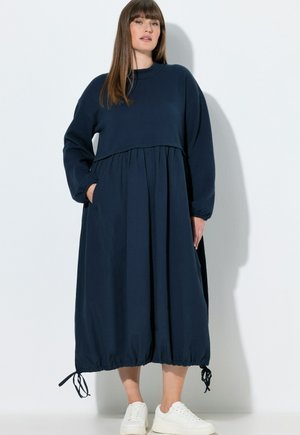 Vrouw met steil bruin haar draagt een losse, lange marineblauwe jurk met aangetrokken zoomtouwen en witte sneakers, staand tegen een witte muur.