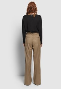 Vrouw met golvend rood haar, gekleed in een zwarte lange mouw top en hoog opgesneden bruine gestreepte wijde broek, die met haar rug naar de kijker staat.