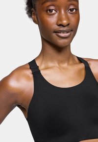 Sostenedor deportivo negro con una textura suave, tirantes anchos y herrajes ajustables. Presenta un cuello redondeado y un diseño minimalista.