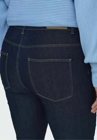 Nära bild av en person som bär mörkblå denimjeans och en ljusblå ribbad tröja, som visar bakfickor och sömnadsdetaljer.