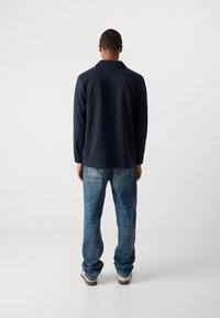 Man standing facing away, wearing a dark long-sleeve shirt, blue jeans, and light-colored sneakers against a plain white background.