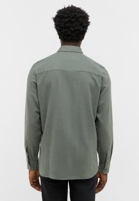 Zielona koszula z długim rękawem, wykonana z miękkiego materiału, o prostym kroju, z kołnierzykiem i zapięciem na guziki przy mankietach. Widoczne subtelne faktury i szczegóły odcinka na plecach.