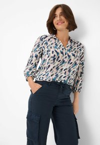 Blouse met een kraag, middenlange mouwen en een veelkleurig abstract patroon in blauw, groen en roze op een lichte achtergrond. Gecombineerd met marineblauwe cargobroek.