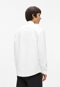 Balta garās piedurknes krekls, izgatavots no gludas auduma, ar klasisku apkakli un pogām pie piedurknēm, ar taisnu griezumu un aizmugurē apdari.