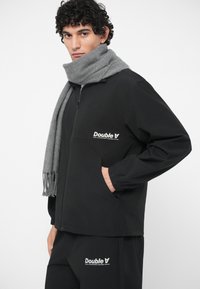 Crna jakna s prednjim patentnim zatvaračem, elastičnim manžetama i bočnim džepovima. Sivi šal s resama. Oboje nosi logo "Double A".