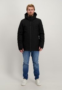 Zwarte geïsoleerde jas met een capuchon, ritssluiting, zijzakken en een gestructureerde stof. Draag het over lichtblauwe jeans en witte sneakers.