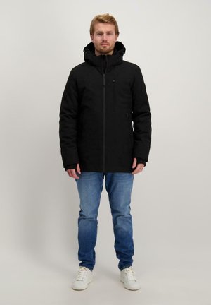 Schwarze isolierte Jacke mit Kapuze, Reißverschluss, seitlichen Taschen und strukturiertem Stoff. Getragen über hellblauen Jeans und weißen Sneakers.