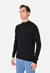 Maglione nero a maglia con collo alto e polsini a coste, caratterizzato da un design aderente e maniche lunghe. Indossato con jeans azzurri chiari.