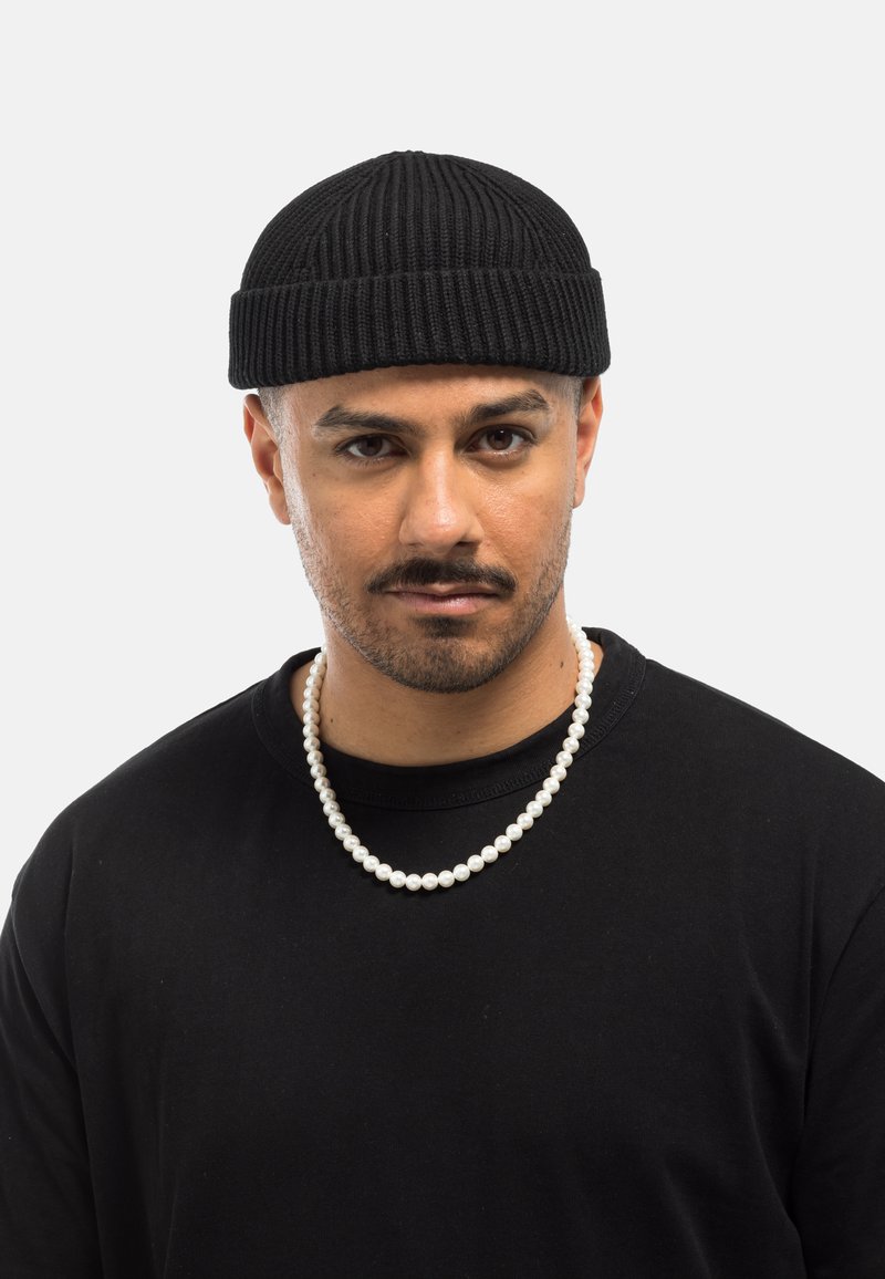 Gorro de punto negro con textura acanalada, combinado con una camiseta negra y un collar de perlas, que presenta cuentas blancas redondas y una superficie suave.