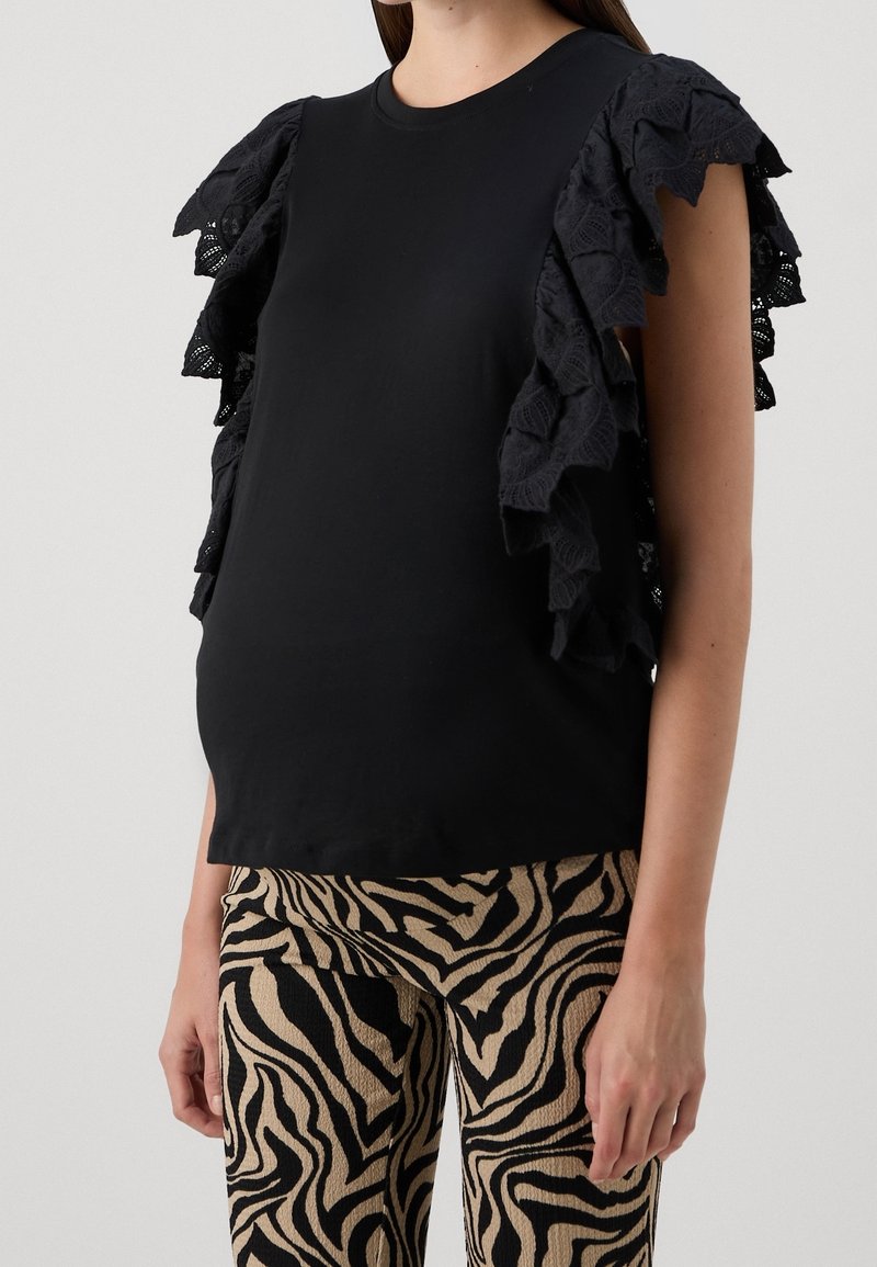 Femme portant un haut noir sans manches avec des manches à volants en dentelle superposée et un pantalon beige à imprimé zèbre noir, debout devant un fond uni.