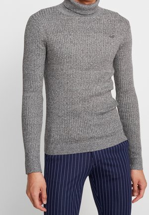 Homme portant un pull moulant gris à col roulé côtelé et un pantalon bleu marine à fines rayures, debout devant un fond blanc.