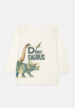 Maglietta a maniche lunghe color crema realizzata in tessuto morbido, con illustrazioni di dinosauri colorati e la scritta "DINO SAURUS" in lettere grasse di colore verde scuro.