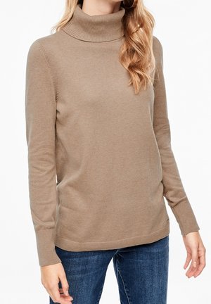 Donna che indossa un maglione beige a collo alto e jeans blu, in piedi con le braccia rilassate lungo i fianchi davanti a uno sfondo bianco.