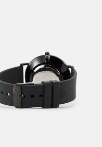 Montre en silicone noir avec un boîtier rond, un bracelet texturé et une boucle en métal. L'arrière présente un design lisse avec des informations sur le modèle.