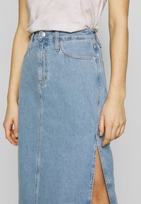 Džínová sukně světle modré barvy s předním rozparkem, zipem a zapínáním na knoflík. Obsahuje klasické prošívání a boční kapsy. Udržitelný materiál.