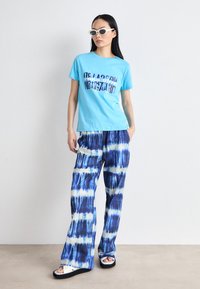 Blue cotton T-shirt with graphic text, paired with tie-dye wide-leg pants in blue and white. Model wears white sandals and sunglasses.