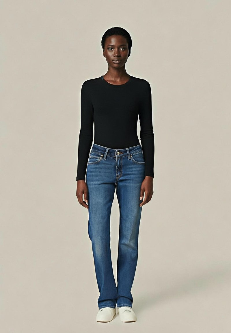 Una donna che indossa un top nero aderente a maniche lunghe, jeans blu a gamba dritta e scarpe bianche senza lacci, in piedi contro uno sfondo beige uniforme.