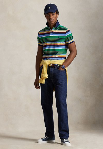 Mann iført marineblå caps, grønnstripet polotrøye, gul genser knyttet rundt livet, mørke jeans, blå klokke og joggesko, stående foran en ensfarget bakgrunn.