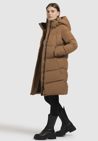 Ruskea puhallettava takki, jossa on pitkä siluetti, huppu ja vetoketjullinen etuosa. Ominaisuuksina suuret tikatut osiot, kaksi sivutaskua ja musta vuori.