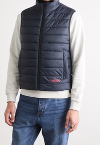 Granatowa kamizelka puchowa z poziomymi przeszyciami, wysokim kołnierzem, zamkiem z przodu oraz kieszeniami bocznymi. Czerwone logo "HUGO" po lewej stronie. Casualowy krój.