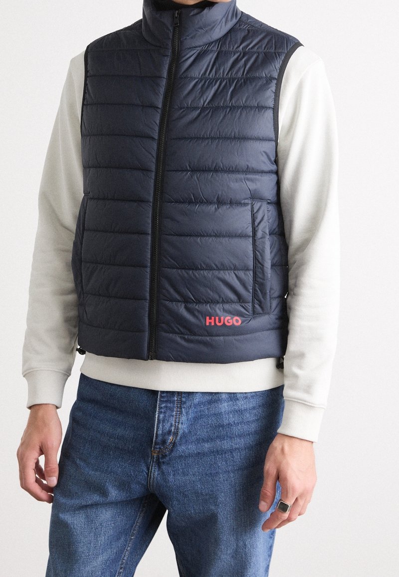 Granatowa kamizelka puchowa z poziomymi przeszyciami, wysokim kołnierzem, zamkiem z przodu oraz kieszeniami bocznymi. Czerwone logo "HUGO" po lewej stronie. Casualowy krój.